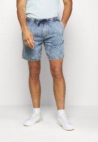 Hombre con pantalones cortos de mezclilla lavados ácidos azul claro, zapatillas blancas, calcetines blancos y una camiseta de color claro, de pie con una mano en el bolsillo.