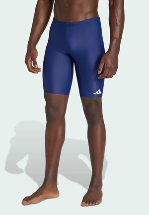 Männliches Model trägt eng anliegende marineblaue Badeshorts mit weißem Adidas-Logo am linken Oberschenkel und steht barfuß auf hellem Hintergrund.