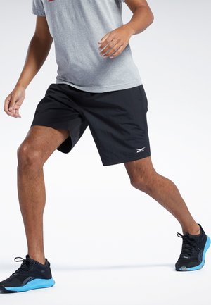 Mustat urheilushortsit, joissa on kevyt kangas, polvipituinen leikkaus ja pieni valkoinen logo. Yhdistetty mustiin kenkiin, joissa on sinisiä yksityiskohtia.