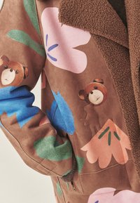 Brązowa kurtka z teksturowanym, puszystym kołnierzem, zdobiona kolorowymi kwiatowymi wzorami i ilustracjami niedźwiedzi w różowym, niebieskim i zielonym kolorze.