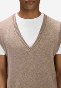 Stickad ärmlös tröja i beige, med djupt V-ringad hals och ribbade kanter. Bärs över en vit T-shirt med rund hals.