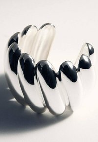 Pulsera de puño de plata pulida con segmentos gruesos, lisos y redondeados que crean una forma circular abierta y flexible sobre una superficie blanca.