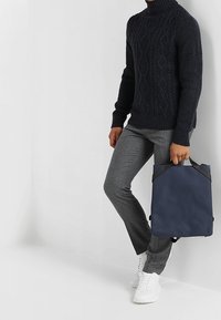 Persona vestita con un maglione a trecce nero e pantaloni grigi, che tiene uno zaino blu navy per la maniglia superiore, in piedi contro un muro bianco.