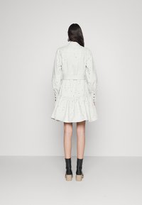 Robe blanche avec manches bouffantes, jupe à volants et ceinture arrière. Présente de petits trous pour la texture. Portée avec des bottines noires.