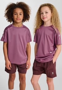 Twee kinderen dragen bijpassende sportoutfits. Beide hebben een paars T-shirt met korte mouwen en donker bordeauxrode korte broeken met witte accenten en logo's.