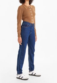 Kvinna klädd i brun ribbad långärmad knäpptröja, högmidjade blå jeans och vita sneakers med svarta ränder, stående.