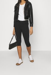 Pantalons noirs coupés, associés à une chemise blanche. Portés avec une veste en cuir noire et des baskets blanches, tenant un sac à main noir.