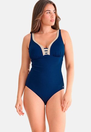 Jonge vrouw in een marineblauw eendelig badpak met beige rand en gestreepte binnenkant, staand tegen een effen achtergrond.