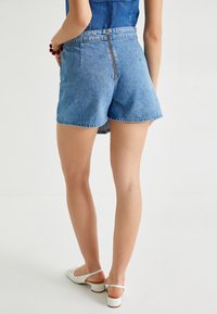 Pantalones cortos de mezclilla en azul claro, con cremallera trasera, dos bolsillos laterales y dobladillo enrollado. Textura suave con diseño clásico de jeans.