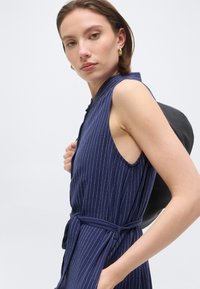 Γυναίκα με κοντά καστανά μαλλιά που φορά φορεμα χωρίς μανίκια σε σκούρο μπλε χρώμα με ρίγες, κρατώντας μαύρη τσάντα στον ώμο, κοιτάζει προς την κάμερα.