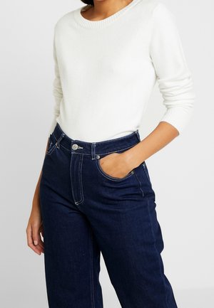 Personne portant un pull blanc à manches longues, rentré dans un jean taille haute bleu foncé, avec une main partiellement dans la poche.