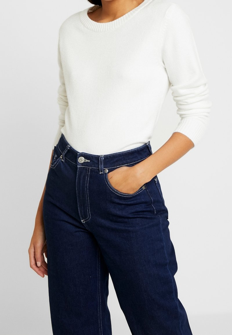 Personne portant un pull blanc à manches longues, rentré dans un jean taille haute bleu foncé, avec une main partiellement dans la poche.
