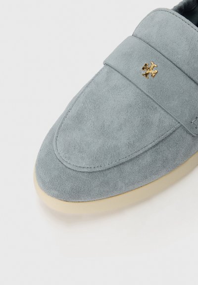 Mocassini in suede grigio con punta arrotondata, caratterizzati da un accento in oro e una suola in gomma di colore chiaro. Texture liscia e design minimalista.
