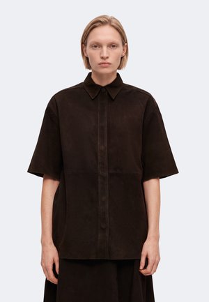 Personne aux cheveux blonds courts portant une chemise noire ample à manches courtes avec un col et des boutons sur le devant.