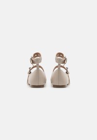 Beige leren ballerina's met ronde neuzen, enkelbandjes met decoratieve gaatjes en een klein strikdetail op de hiel, weergegeven tegen een witte achtergrond.