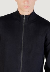 Joven vestido con un suéter negro con cremallera y un cuello de pie acanalado, visible desde el cuello hasta la mitad del torso, contra un fondo neutro.