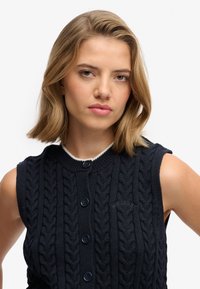 Jersey de punto sin mangas de color azul marino con diseño trenzado, cuello en contraste blanco y cuatro botones oscuros. Incluye el logo de la marca bordado en el pecho.