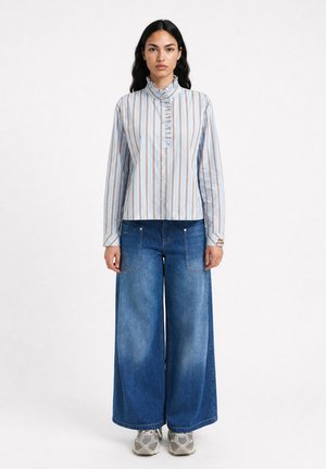 Woman stands facing forward wearing light blue and brown striped long-sleeve shirt with ruffled collar and wide-leg blue jeans with sneakers.