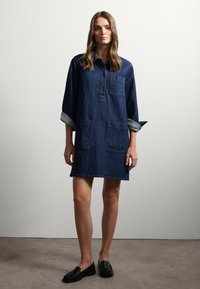 Donkerblauwe denim jurk met lange mouwen, een knoopsluiting aan de voorkant en twee grote voorzakken. Zachte textuur met opgerolde mouwuiteinden.