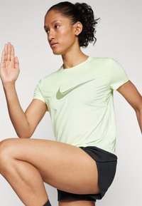 Kortermet, lysegrønn treningstrøye med et reflekterende Nike-logo. Brukt sammen med svarte shorts, som viser teksturert hud og en hevet arm.