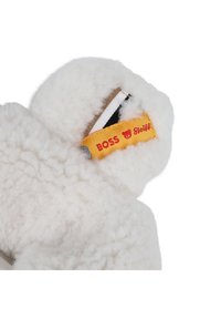 Bílá kozačka z beránkového kožíšku s měkkou texturou a zakřiveným designem. Na botě je žlutá visačka s červeným textem, označená značkami 'BOSS' a 'Steiff'.
