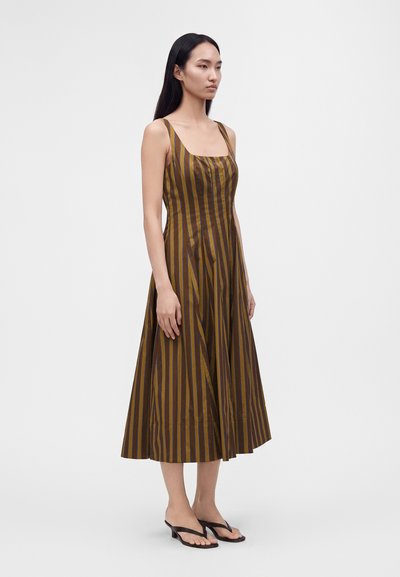 Femme debout portant une robe midi sans manches à rayures verticales marron et jaune avec un décolleté carré et des sandales noires à talons.