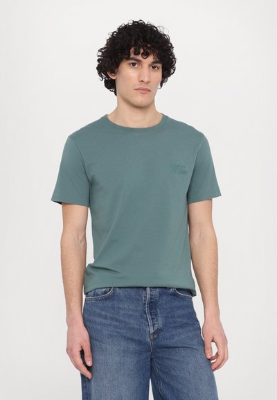 T-shirt en coton vert avec un col rond et des manches courtes, arborant un petit logo ton sur ton sur la poitrine, associé à un jean en denim bleu.