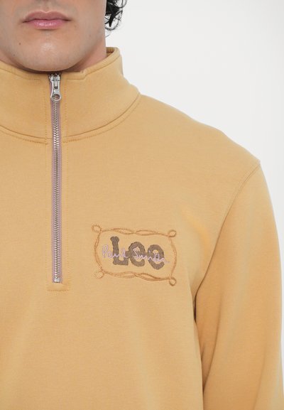 Pull en polaire zippé jaune avec col haut, arborant un logo brodé en marron et rose clair sur la poitrine. Texture lisse et douce.