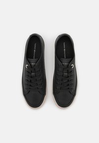 Černé kožené tenisky s texturovaným povrchem, plochými tkaničkami a hnědou gumovou podešví. Disponují malým kovovým akcentem poblíž otvůrků na tkaničky.