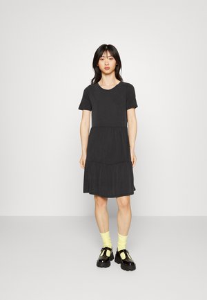 Vestido preto de manga curta com um design em camadas. Tecido suave, decote redondo e um corte descontraído. Usado com meias amarelas e sapatos pretos robustos.