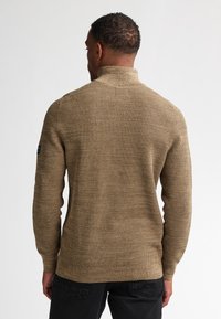 Neulepusero beige-ruskeana, jossa on korkea kaulus ja teksturoitu kangas. Pitkät hihat ja hihansuut on käännetty, vasemmassa hihassa logo-tyyli.