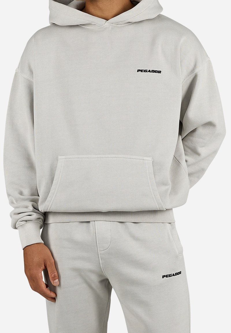 Homme portant un sweat à capuche gris clair et un pantalon de survêtement assorti avec de petits logos noirs "PEGADOR" sur la poitrine et la cuisse.