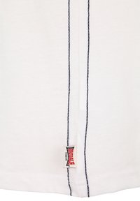 Balta medžiaga su dviem vertikaliomis tamsiai mėlynos spalvos siūtomis siūlėmis ir mažu stačiakampiu „Lonsdale London“ ženklu netoli apatinio krašto.