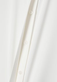 Weiße Bluse aus glattem Stoff, mit Knopfverschluss und einem schlichten Design mit subtiler Textur. Sichtbare Knöpfe auf einer Seite.