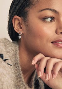 Pendientes plateados en forma de corazón con un diseño recortado y un colgante de corazón texturizado. La modelo lleva un suéter de punto suave.