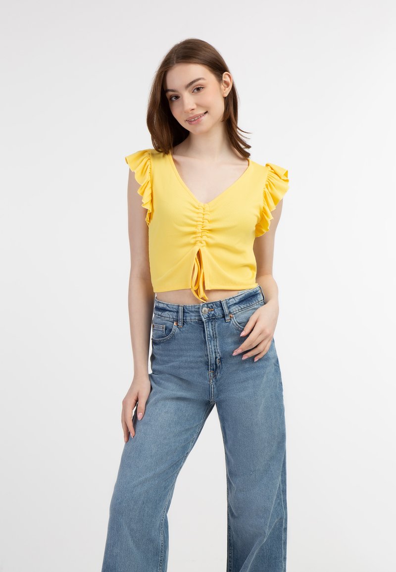 Gul ribbet crop top med flæser på ærmerne og en frontsløjfe, parret med højtaljede lyseblå vide jeans.