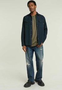 Camicia a bottoni blu scuro, maglietta verde oliva, jeans blu strappati, scarpe nere. Caratteristiche includono due tasche sul petto e segni di usura sui jeans.