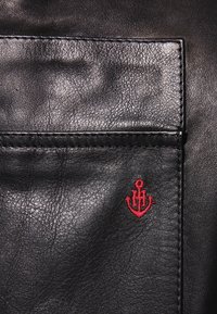 Zwart leren kledingstuk met een steekzak en een klein leren ankerlogo in het rood geborduurd. De textuur is glad met zichtbare nerf.