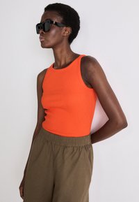 Un maiou portocaliu cu pliuri, asociat cu pantaloni lejeri de culoare verde măslin. Modelul poartă ochelari de soare rectangulare negri.