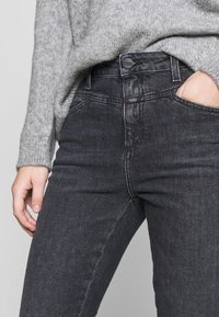 Jeans gris oscuro de corte slim, con cierre de botón metálico, ligero desgaste y bolsillos frontales. Llevado con un suéter de punto gris claro.