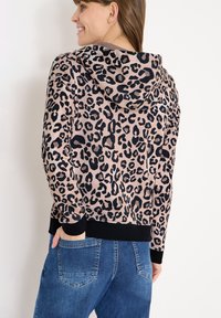 Kapuzenpullover mit Leopardenmuster aus weichem rosa Stoff mit schwarzen Flecken, ausgestattet mit einer Kapuze und schwarzem geripptem Saum. Kombiniert mit blauen Denim-Jeans.