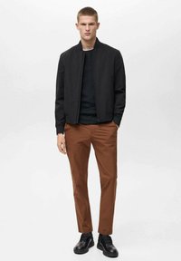 Musta pommikauppias, tumma villapaita, ruskeat housut ja mustat nauhaliuskat. Siistejä linjoja, istuva muotoilu, jossa on sileä kangasrakennete.