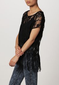 Svart spetsblus med blommönster, lös passform, korta ärmar och fransdetaljer vid fållen. Kombinerad med blå denimjeans.