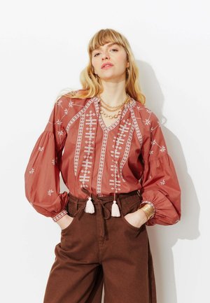 Femme aux cheveux blonds portant un chemisier brodé couleur rouille à manches bouffantes et un pantalon taille haute marron, les mains dans les poches.