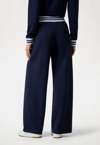 Marine-blauwe wijde broek gemaakt van gladde stof, met een gestreepte tailleband en boorden. Witte sneakers complementeren de outfit.