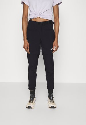 Persoon die zwarte afritsbroek met riem draagt, wit kort mouwen shirt met knoop, en beige sportschoenen, staand tegen een witte achtergrond.