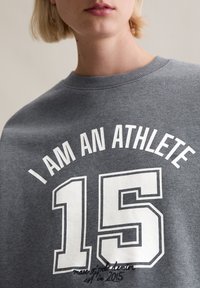Szara bluza z dużym białym napisem "JESTEM SPORTOWCEM" ponad numerem "15". Zawiera detal szycia u dołu.