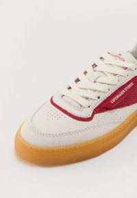 Sneaker med en kremfarget overdel, røde lærdetaljer og perforert design. Har en gummiguttersåle og hvite snøringer.