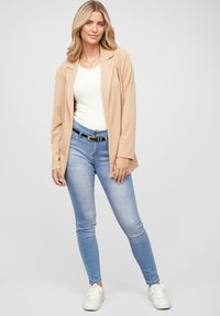 Beige blézer egy fehér felső fölött, világoskék skinny farmerrel és fehér sportcipővel párosítva. Az öltözethez fekete öv és arany kiegészítők tartoznak.