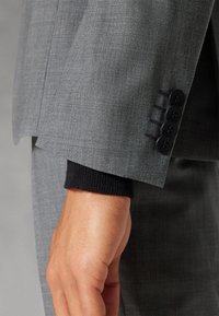 Siva jakna s odrezak od teksturirane tkanine, s crnim gumbima kao detaljem i crnim pletenim manžetama. Vidljivo detaljno šivanje u blizini gumba.